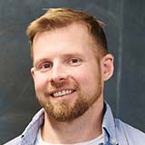 A bearded man with short light brown hair smiles while facing the camera, wearing a light blue collared shirt. The background is a dark, out-of-focus surface.